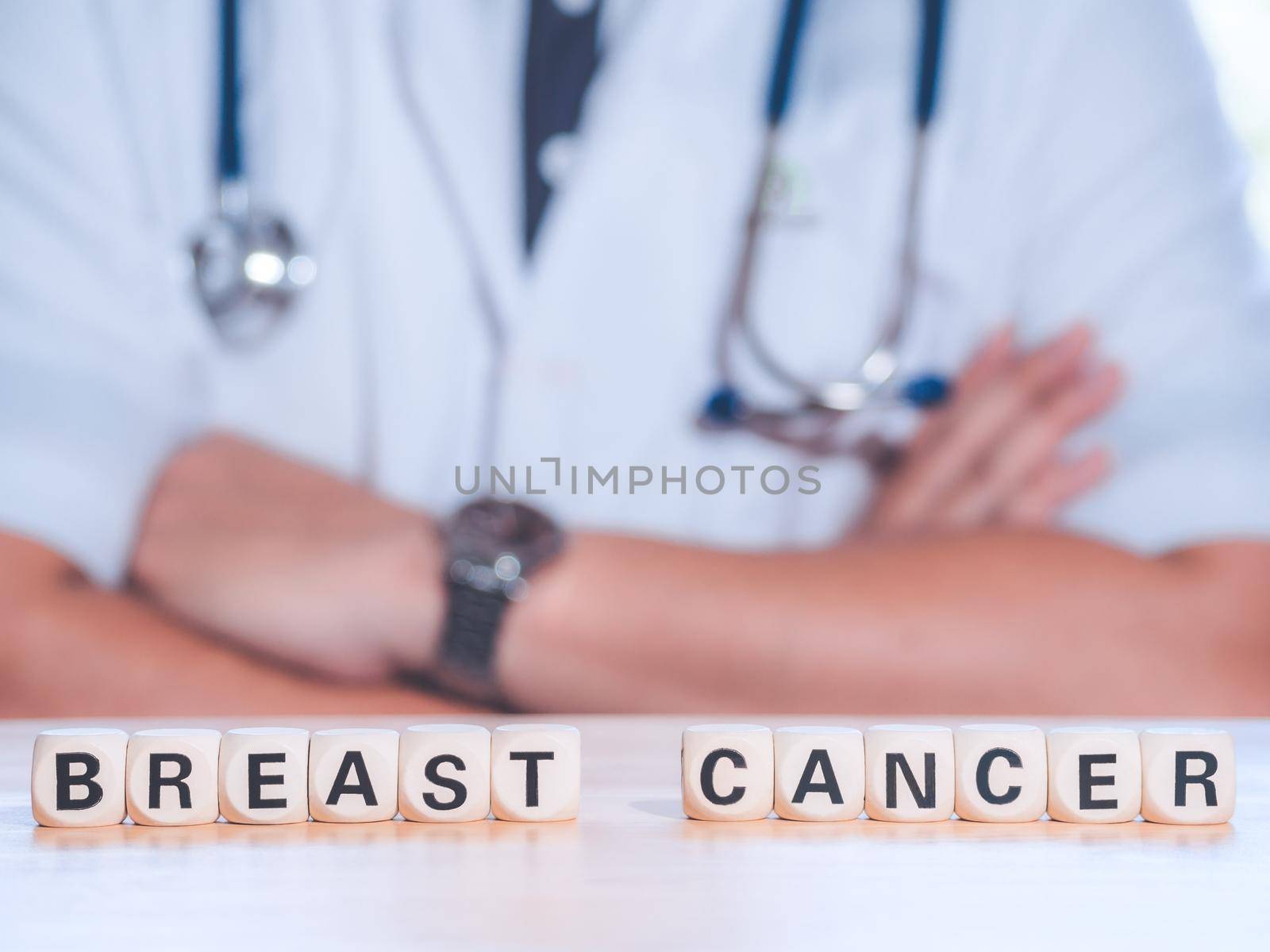 Closeup wooden block written BREAST CANCER on table against professional male doctor with stethoscope. Physician ready to examine patient. Medical and patient care concept. by Chakreeyarut