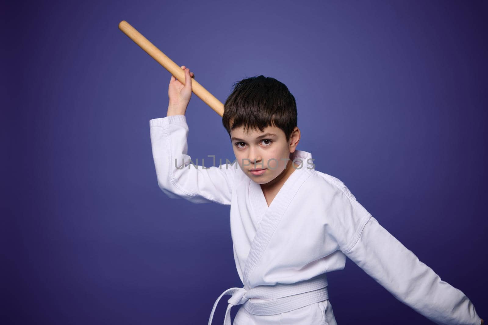 Confident charming European aikido wrestler poses with wooden weapon during Oriental martial arts practice, isolated on violet background, copy ad space for text. Oriental martial arts practice