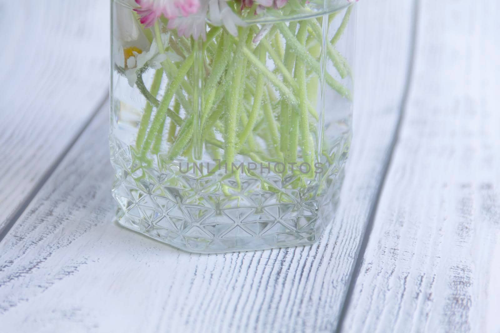 A bouquet of fresh flowers, daisies on green stems stand in a glass with clean water on a gray, wooden background.