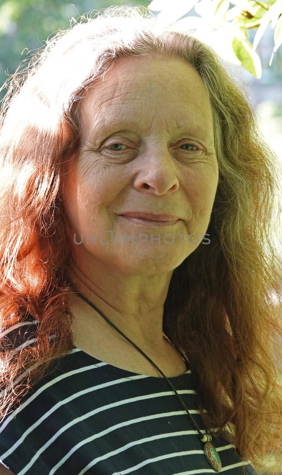 Portrait of a smiling elderly lady without make-up. Her hair is mixed coloured: grey and red. She has wrinkles in her face and is wearing a striped shirt.