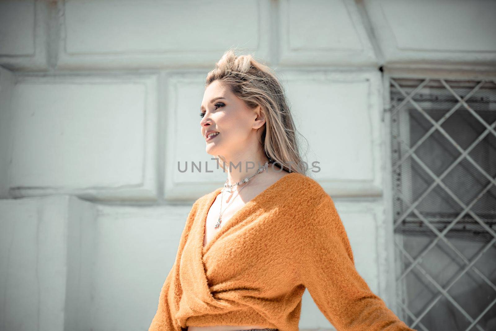 A middle-aged woman looks like a good blonde with curly beautiful hair and makeup on the background of the building. She is wearing a yellow sweater