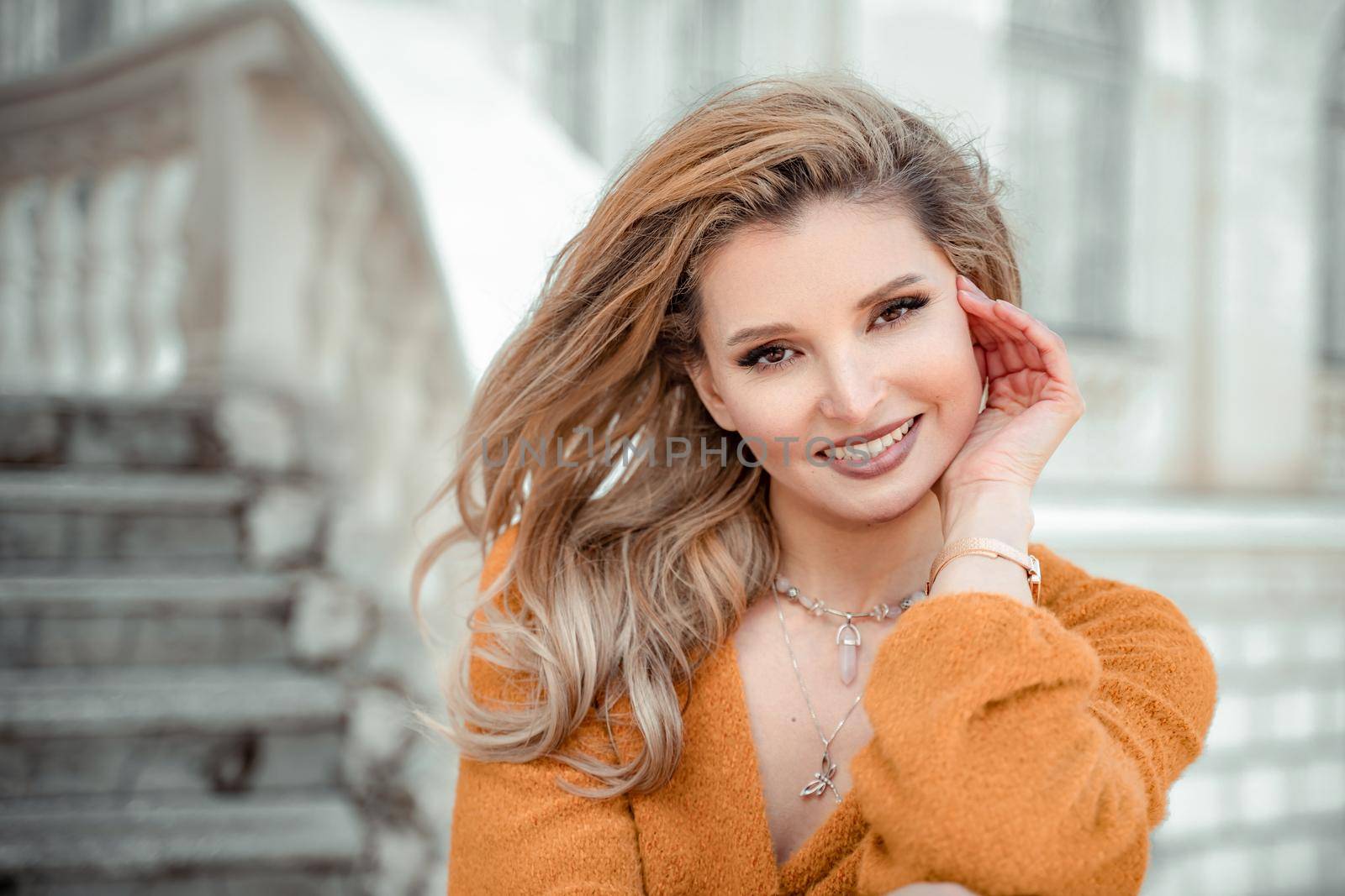 A middle-aged woman looks like a good blonde with curly beautiful hair and makeup on the background of the building. She is wearing a yellow sweater and looking at the camera. by Matiunina