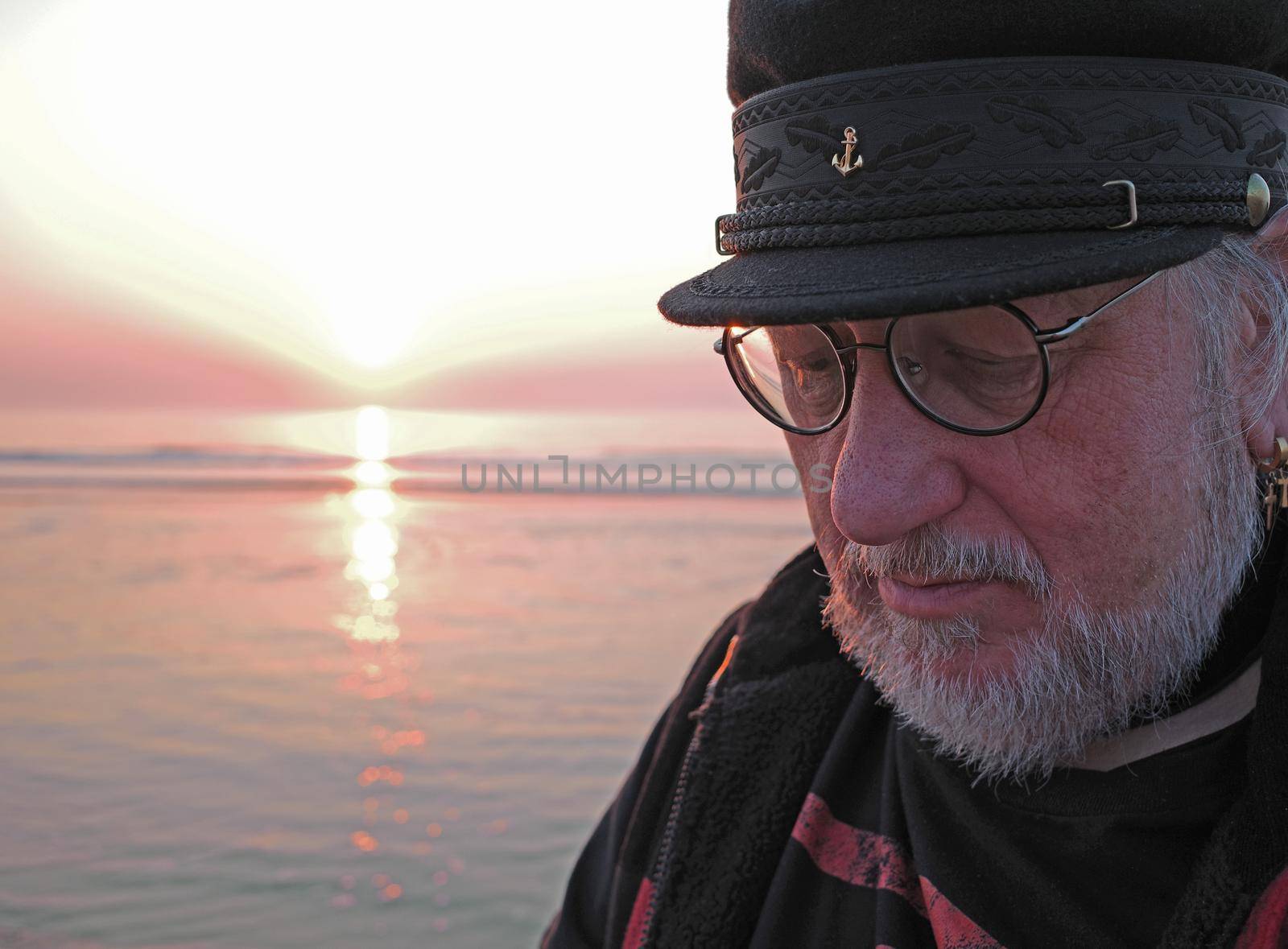 An elderly man is looking thoughtfully down. He is standing in front of the sea. The sun is going down at the horizon. The man has a white beard and a sailor cap on his head