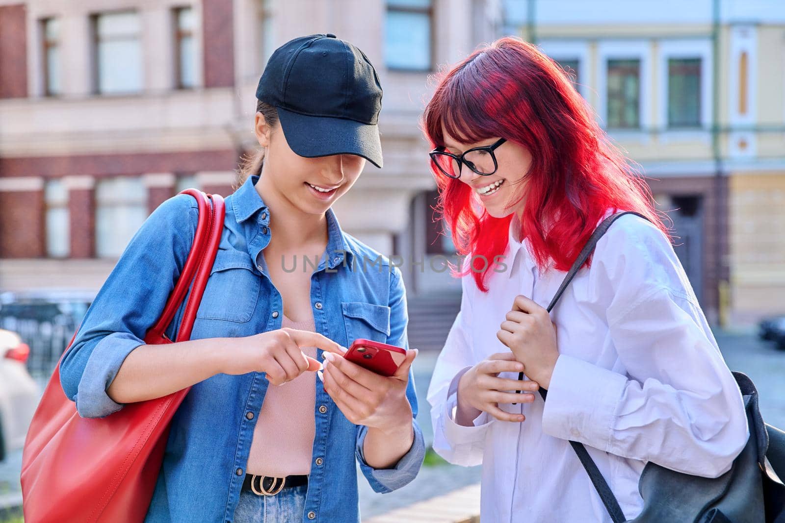 Two teenage females talking, looking into smartphone, outdoor on city street by VH-studio