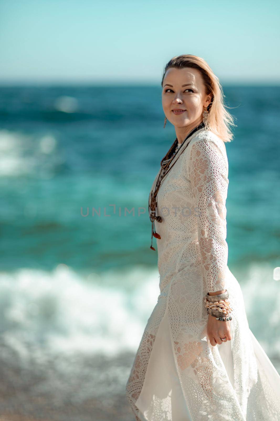 Middle aged woman looks good with blond hair, boho style in white long dress on the beach decorations on her neck and arms