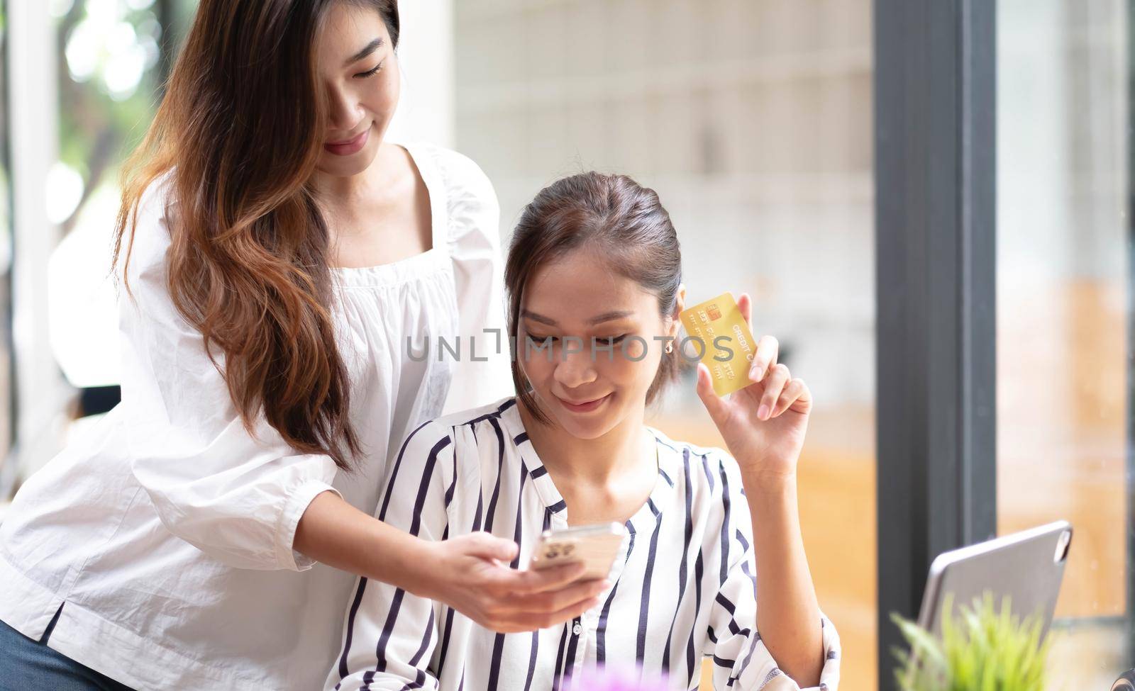 Asian two woman friend enjoy shopping online at cafe. Beautiful lesbian couple sit on sofa, holding credit card to make e bank online payment after use phone purchase goods in web store by wichayada