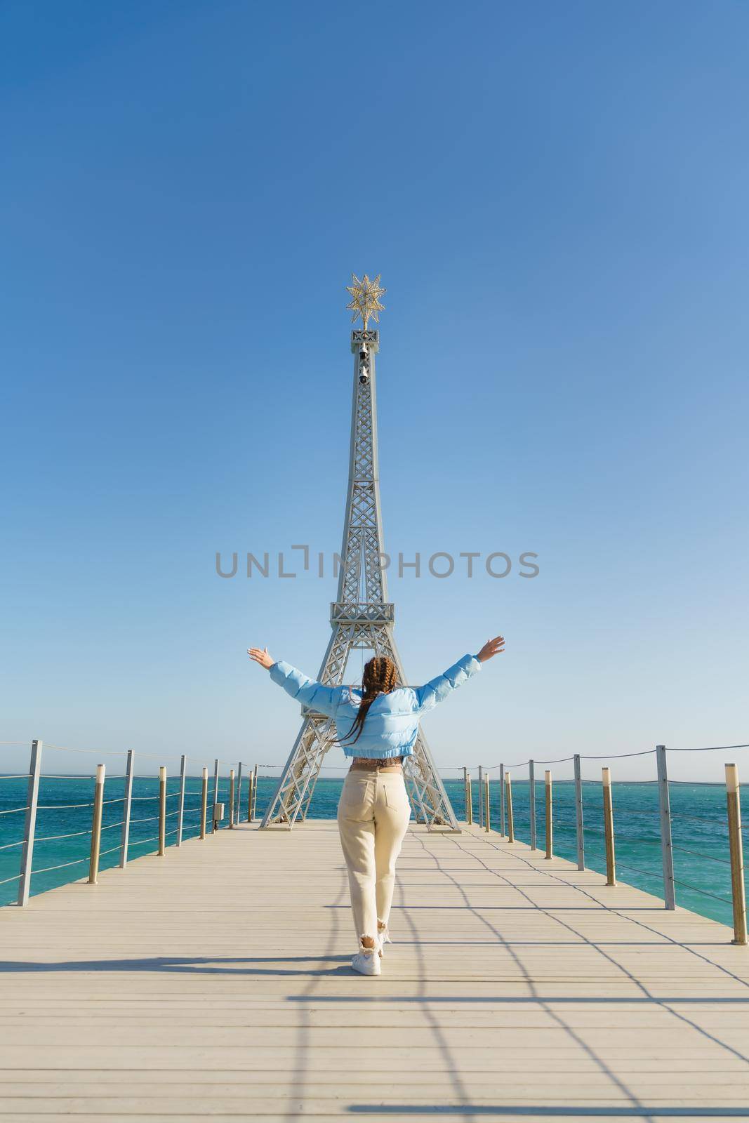 Large model of the Eiffel Tower on the beach. A woman walks along the pier towards the tower, wearing a blue jacket and white jeans. by Matiunina