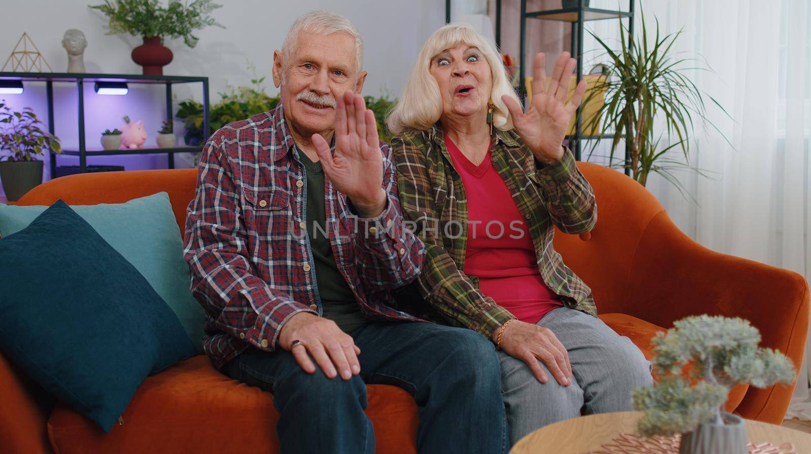 Senior elderly family grandparents man woman smiling friendly at camera and waving hands gesturing hello, hi, greeting or goodbye, welcoming with hospitable expression. Grandmother grandfather at home