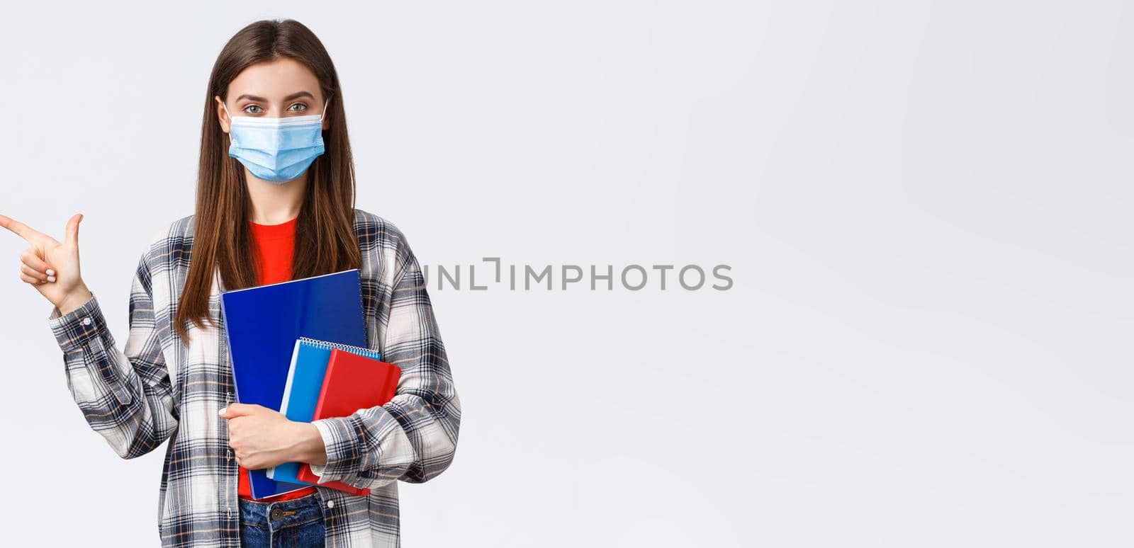 Coronavirus pandemic, covid-19 education, and back to school concept. Young pretty female student in medical mask with notebooks, pointing finger left, showing university info.