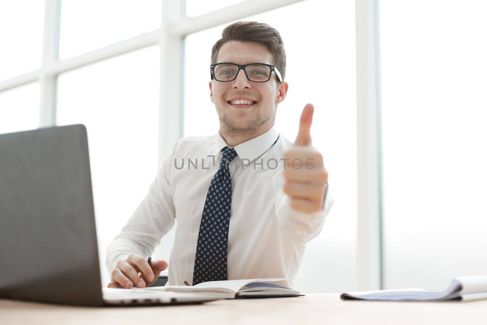 Cheerful young businessman standing and showing thumbs up in office