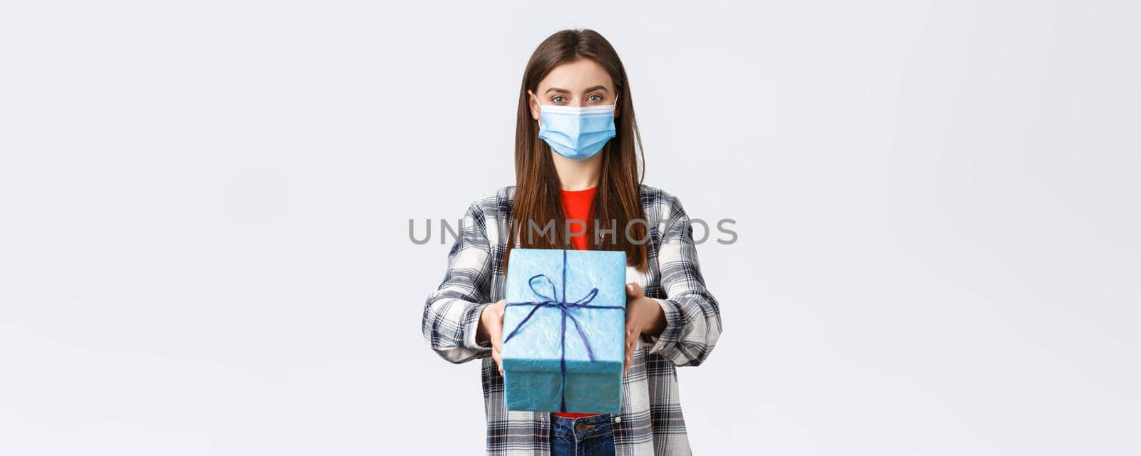 Covid-19, lifestyle, holidays and celebration concept. Cheerful young girl in medical mask stretch hand, handing birthday gift to you, smiling, congratulate with b-day, white background.