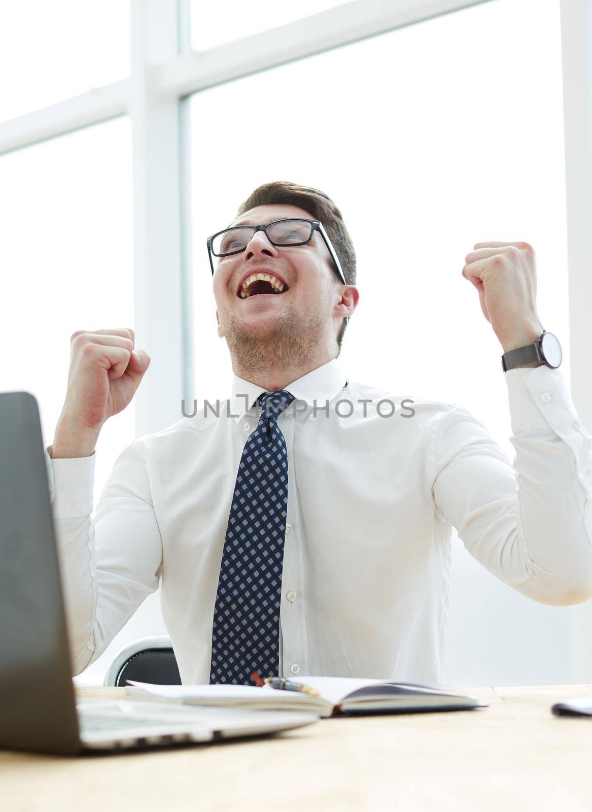 Happy young businessman raising hands in front of laptop at office desk by Prosto