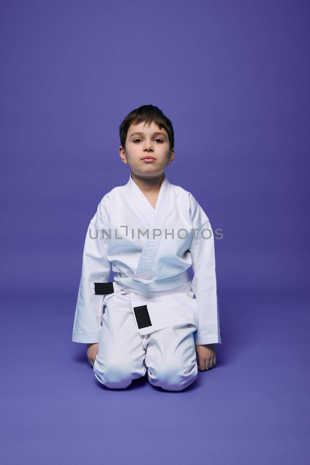 Confident portrait of a handsome child boy wearing kimono and sitting in aikido stance against purple background with copy space for advertising text. Oriental martial arts concept