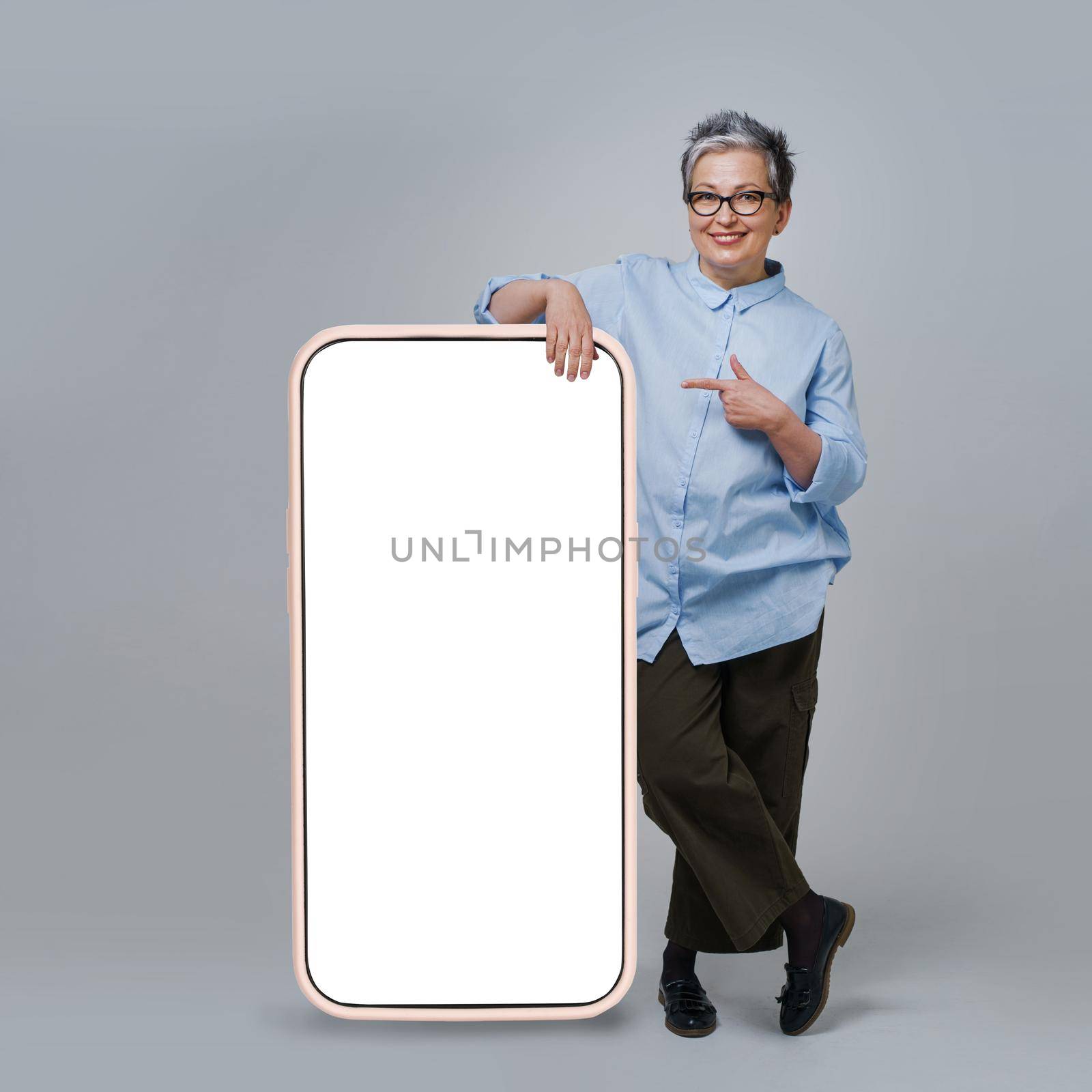 Mature grey hair woman pointing finger on screen of giant, huge smartphone with white, blank screen standing leaned on wearing casual isolated on white background. Free space mock up.