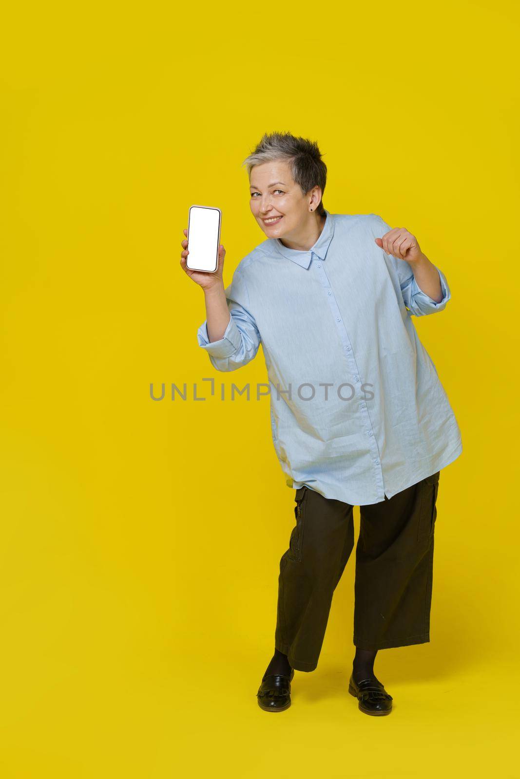 Playful mature grey haired woman smiling holding smartphone showing it white screen. Pretty woman in blue blouse isolated on yellow background. Mobile app advertising. Copy space.