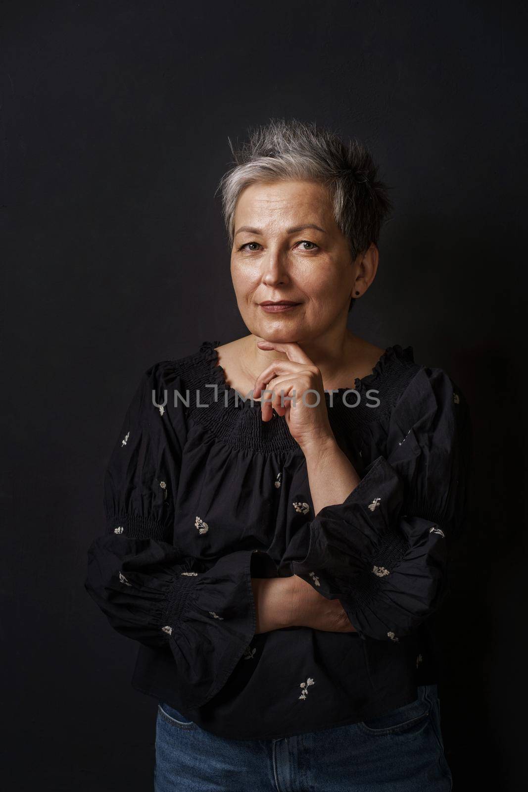 Gorgeous mature businesswoman with grey hair posing leaned chin on her hand isolated on black background. Graying elegant woman with short hair. Image of successful mature woman. Aged beauty.