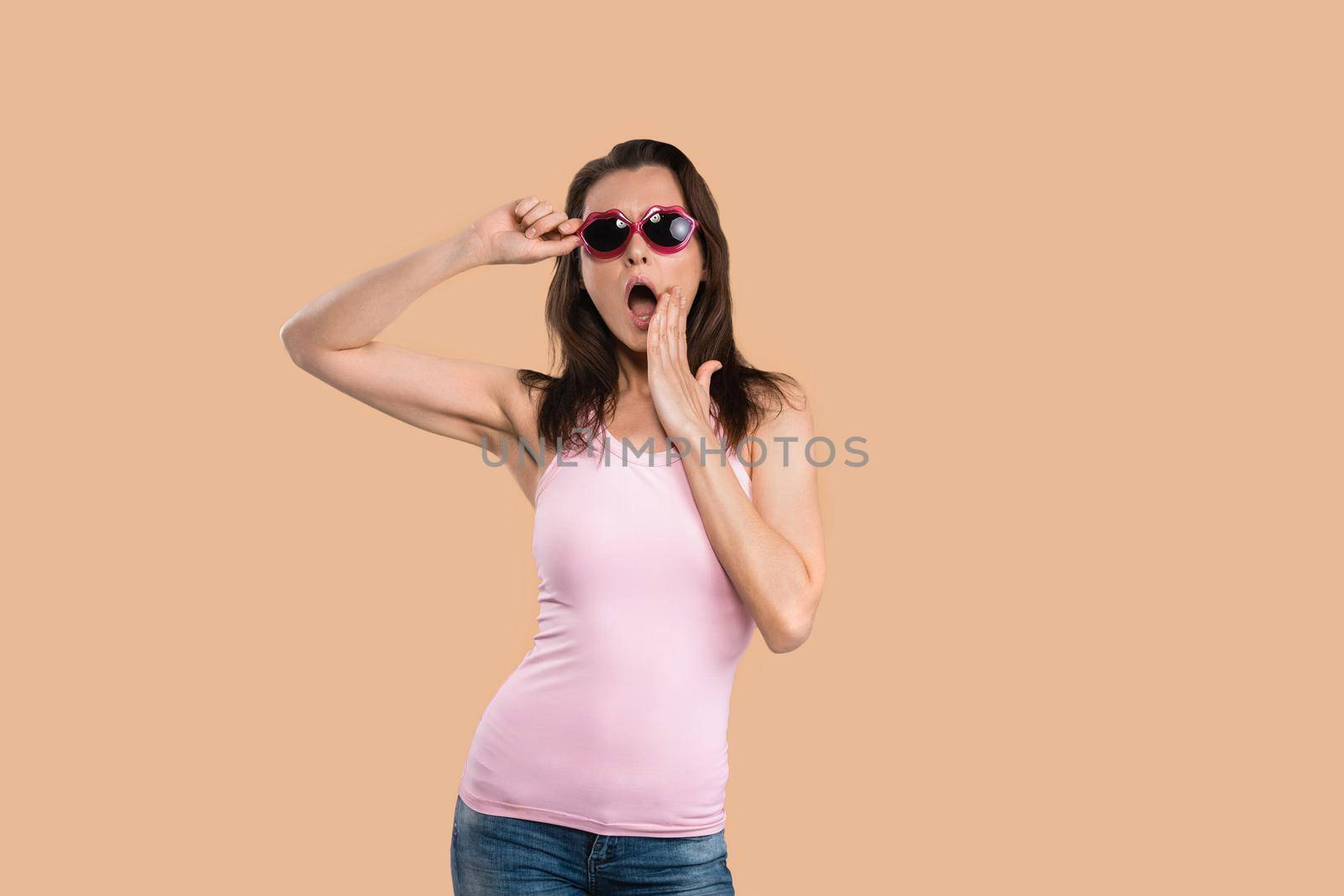 Shocked or wow passionate young woman in sunglasses kiss shaped gesturing oh my god, wearing sleeveless t-shirt posing in studio isolated on beige background.