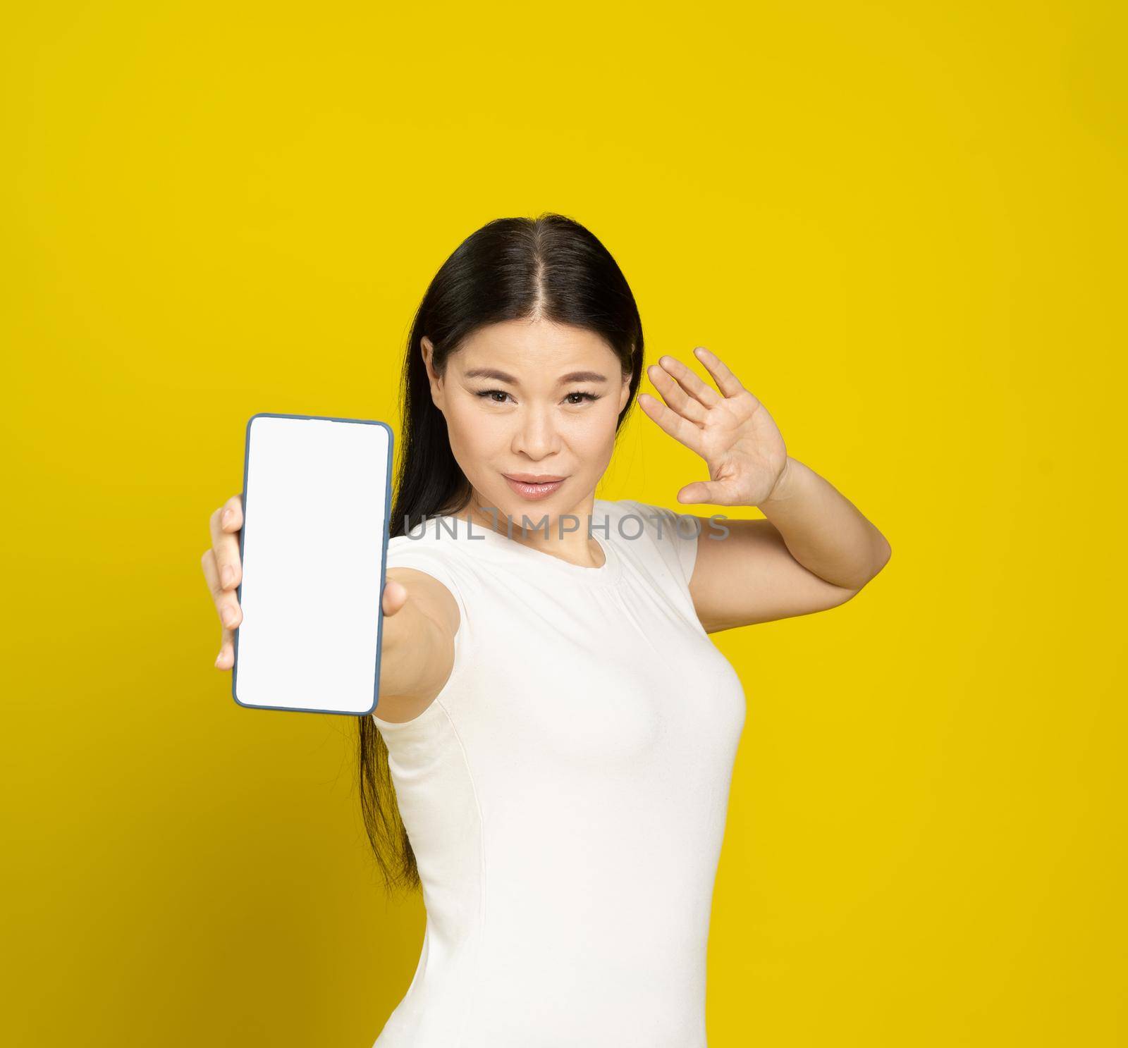 Confident beautiful asian woman 40s smiling holding smartphone showing blank screen mobile app advertisement and smile looking at camera isolated on yellow background. Product placement.