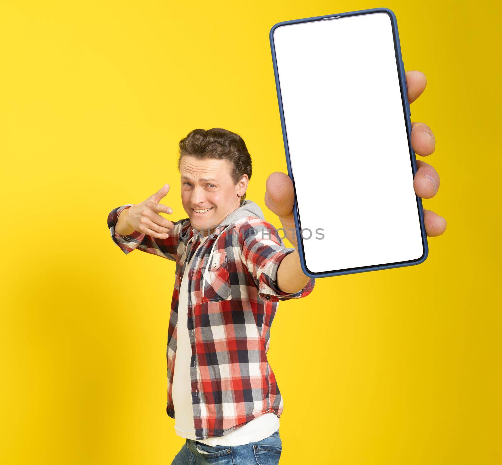 Happy to win handsome man pointing at smartphone with white empty screen, wearing red plaid shirt and jeans cellphone display mock up isolated on yellow background. Mobile app advertisement by LipikStockMedia