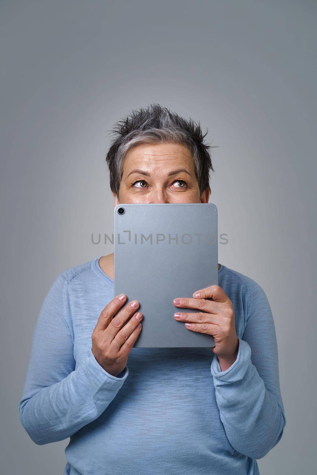 Mature grey haired woman hide face smiling shy behind smartphone or modern gadget working or shopping online or checking on social media. Pretty woman in blue blouse isolated on yellow background by LipikStockMedia