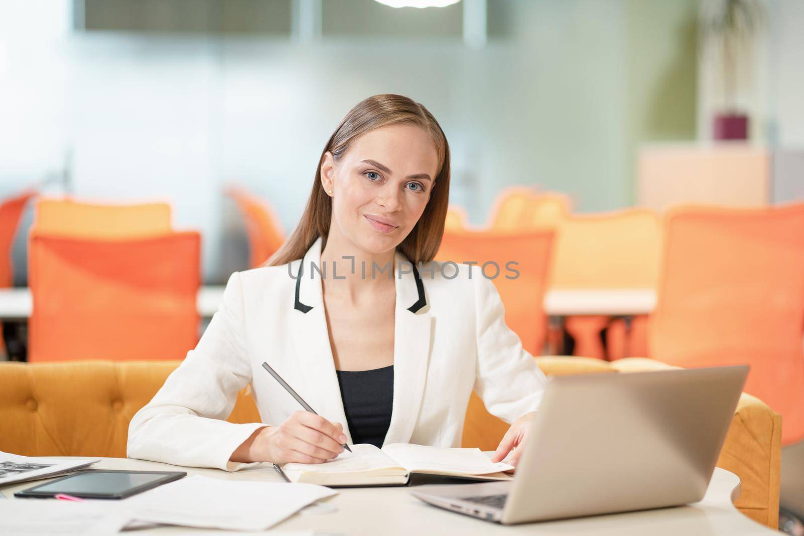 Young business woman in white jacket or suit at the desk with a laptop, looking at camera and making notes. Blonde model female with blue eyes posing in business suit in the office. Business concept by LipikStockMedia