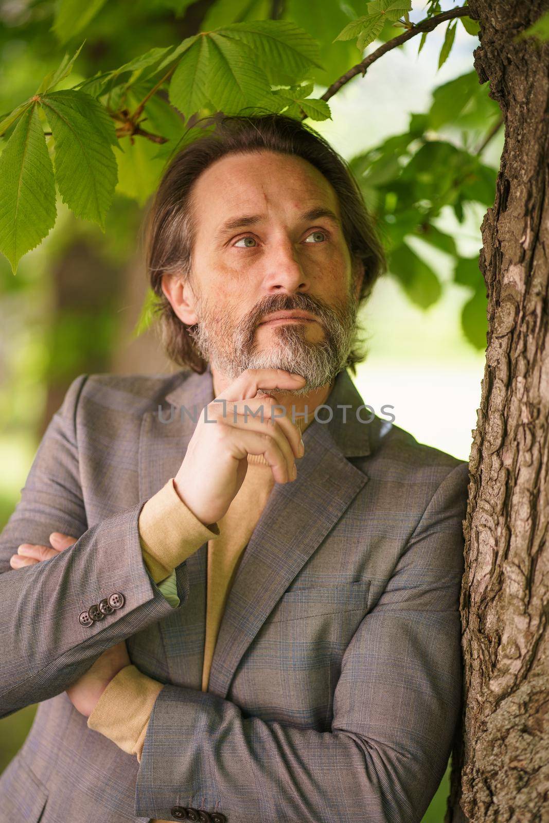 Mature handsome man with grey beard looking up leaning on tree wearing casual grey jacket. Life after 40 years concept, problems and depression. Middle age crisis.