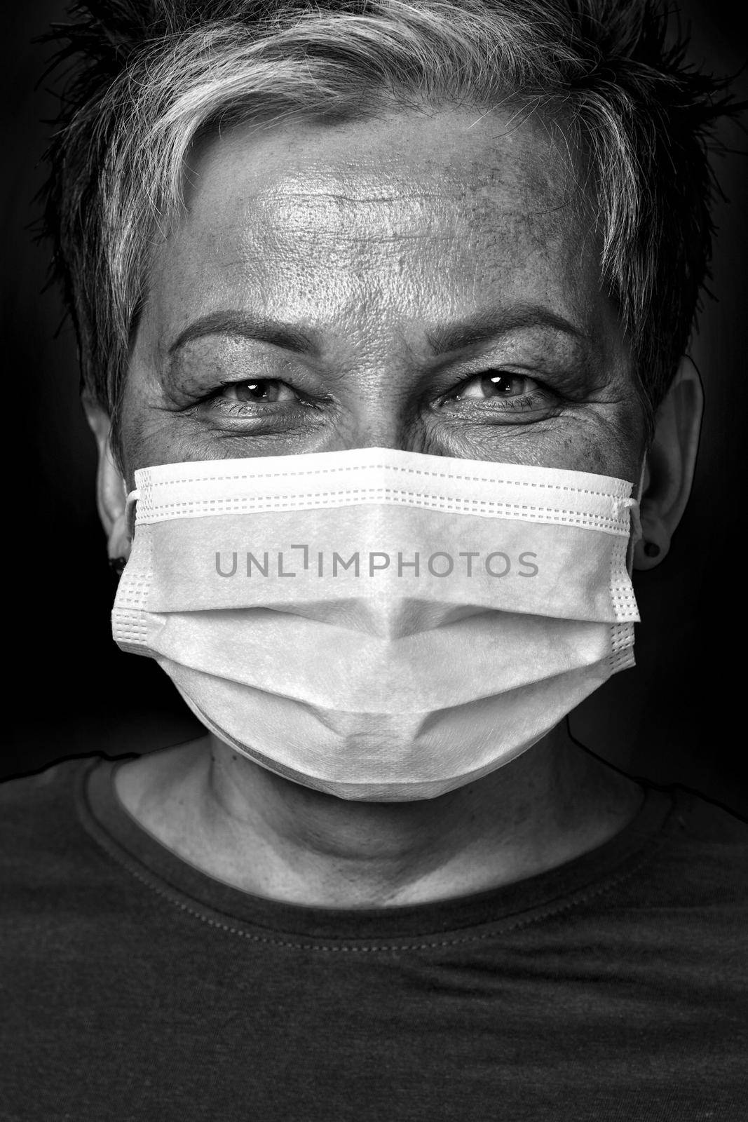 Black and white photo. Happy eyes smiling grey short haired doctor woman wearing medical face mask looking at camera wearing green blouse isolated on yellow background by LipikStockMedia