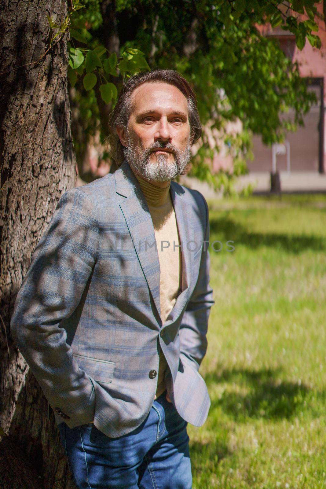 Leaned on tree mature handsome man with grey beard looking at camera wearing casual grey jacket and blue jeans. Life after 40 years concept, problems and depression. Middle age crisis.