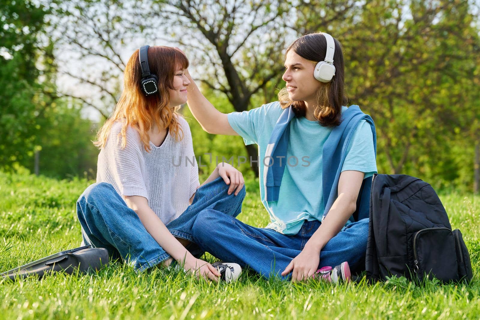 Couple of friends guy and girl 17, 18 years old sitting on grass. Students in headphones with backpacks sitting on campus lawn, talking and laughing. Teens, youth, friendship, education, young people