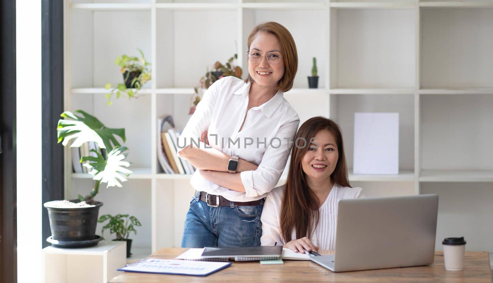 Two middle-aged women are looking at the camera while sitting on a desk in the office. female executives working in the office discussing documents by wichayada