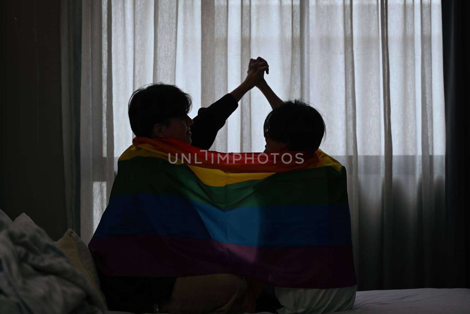Silhouette of gay couple expressing their love to each other under pride flag. LGBT, pride, relationships and equality concept by prathanchorruangsak