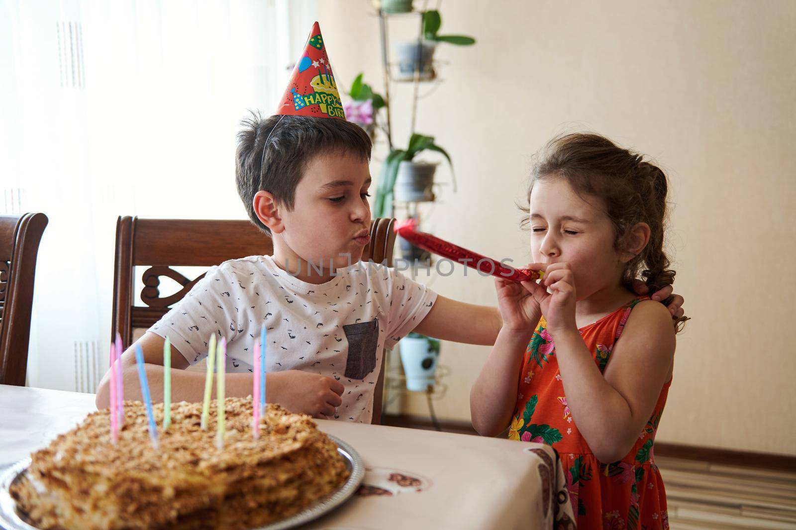 Charming adorable Caucasian kids, boy and girl, brother and sister celebrating birthday party at home. Handmade baked birthday cake with colorful candles on a kitchen table.