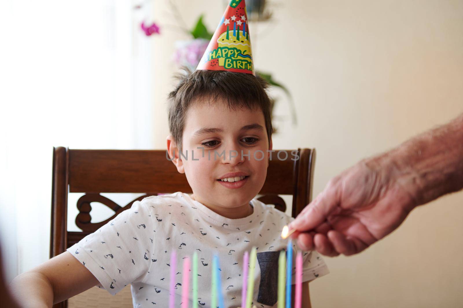 Adorable Caucasian teenage boy looking at grandfather's hand lighting colorful candles on his birthday cake