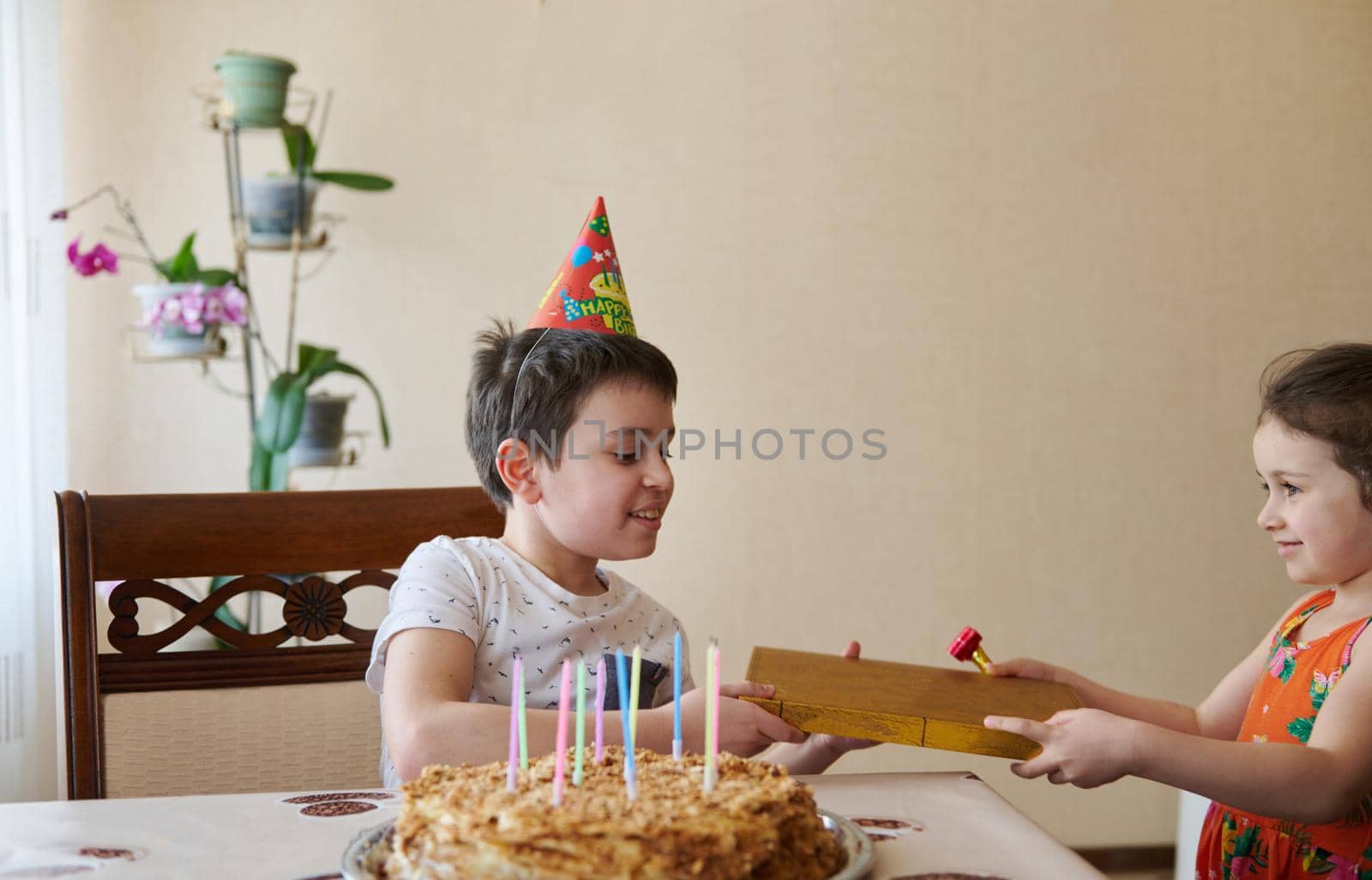 Charming Caucasian boy receives a gift from his younger sister for his 10th birthday, sits at the table with a birthday cake and candles. Birthday, anniversary and event celebration concept by artgf