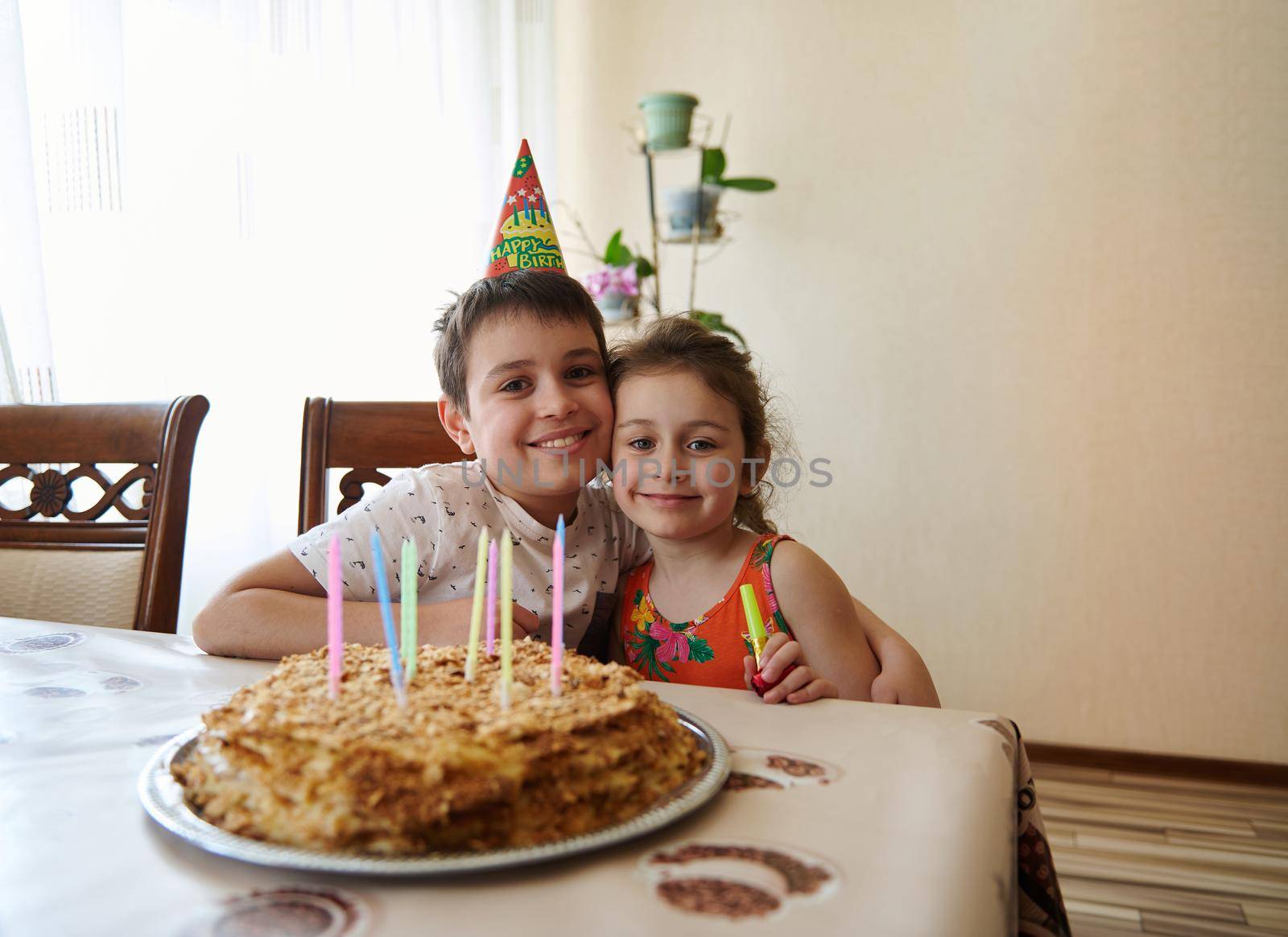 Portrait of two adorable cute Caucasian children, brother and sister, sitting at the table with a birthday cake decorated with long colorful candles. Anniversary and birthday party concept