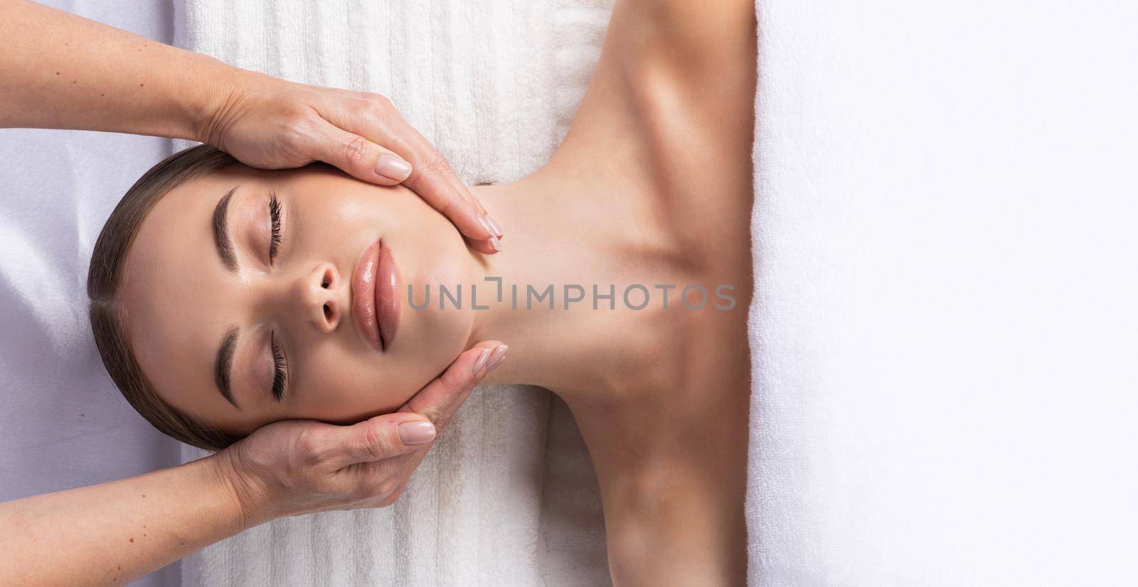 Releasing stress, top view of beautiful young woman lying on back while massage therapist massaging her face, white towel, copy space for text