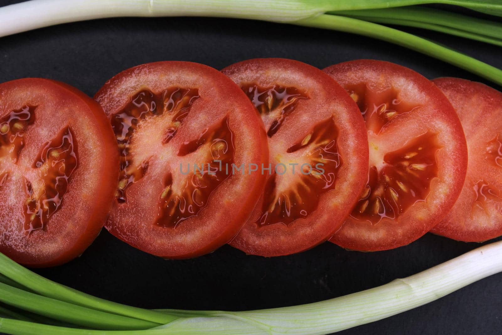Tomato slices in a frame of green onions close-up on a black background. The concept of healthy eating..