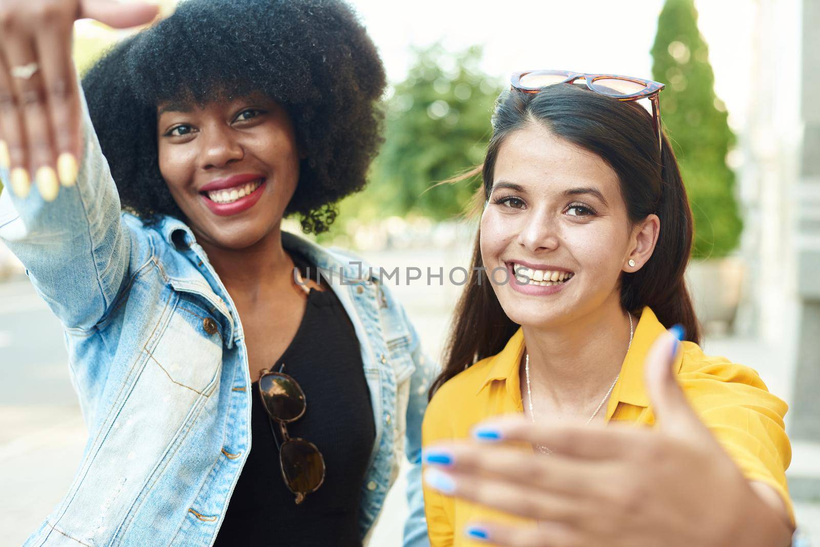 Two beautiful girls of different nationalities show the photo frame with their hands and smile at the camera. Young girlfriends take a selfie on a city street.