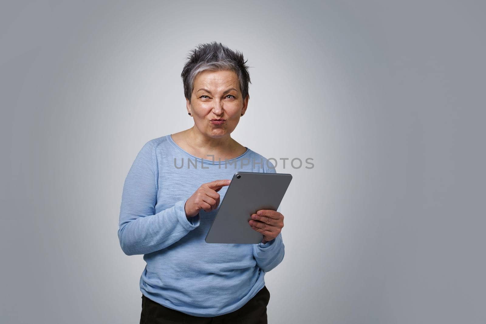 Playful grey haired mature woman with digital tablet working or checking on social media. Pretty woman in 50s in blue blouse isolated on white background. Older people and technologies.