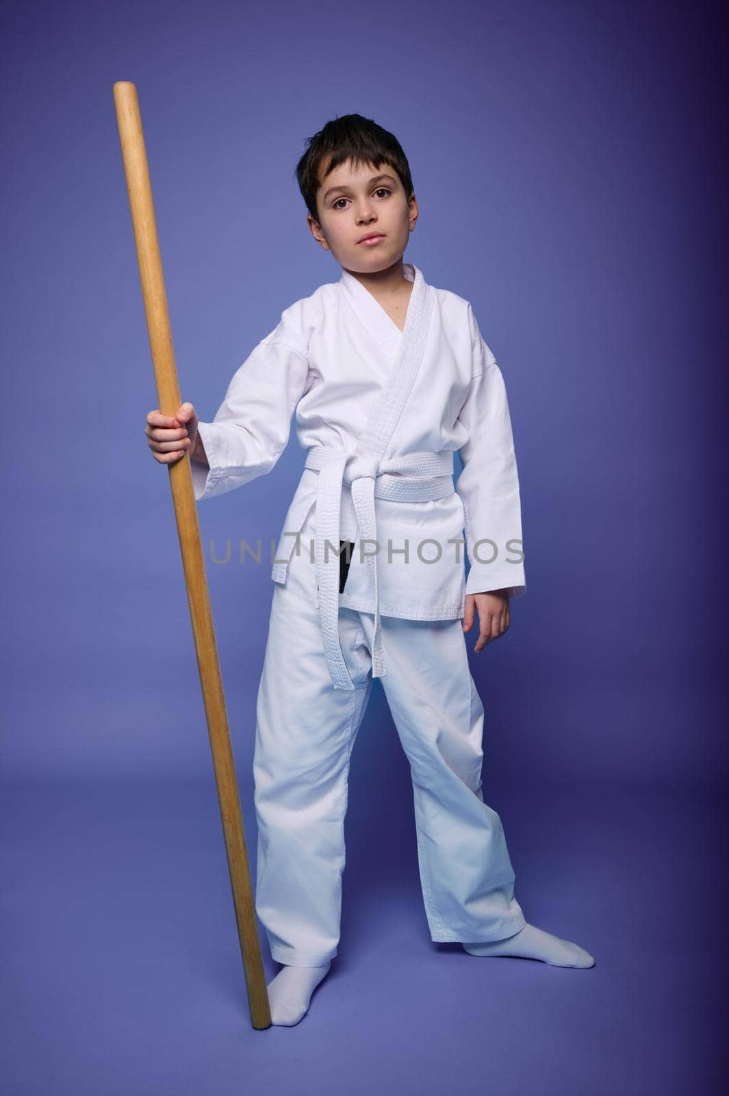Handsome confident 10 tears old child, European teenage boy in white kimono with wooden weapon- bokken standing against purple background with copy ad space. Oriental martial arts concept, Aikido
