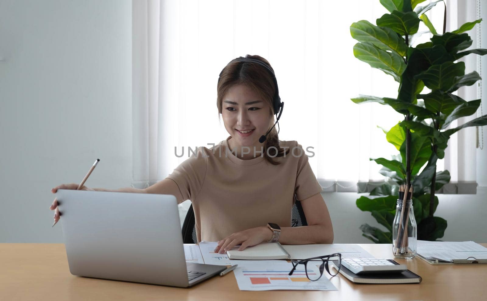 Asian businesswoman talking to colleague team in video call conference writing note on book with smile face. woman using computer laptop and headphone for online meeting. Smart working from home concept. by wichayada