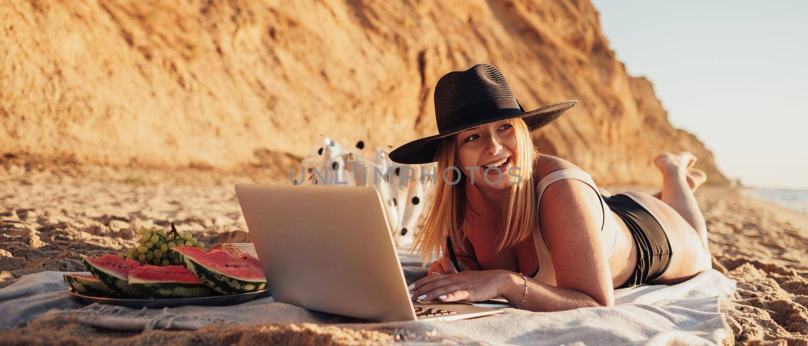 Smiling Young Woman in Swimsuit and Hat Looking Away While Using Laptop on Vacation by Sea