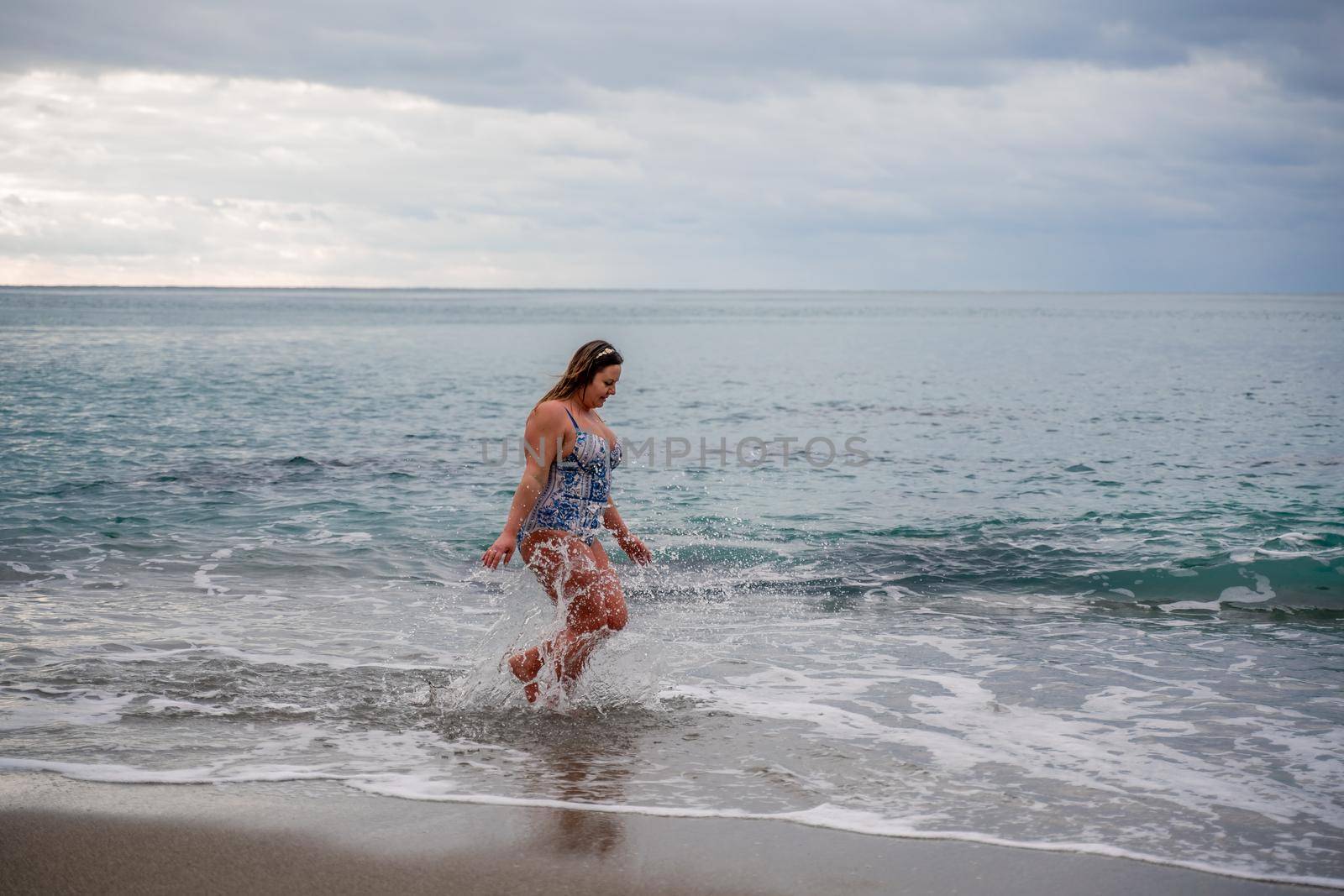 A plump woman in a bathing suit enters the water during the surf. Alone on the beach, Gray sky in the clouds, swimming in winter