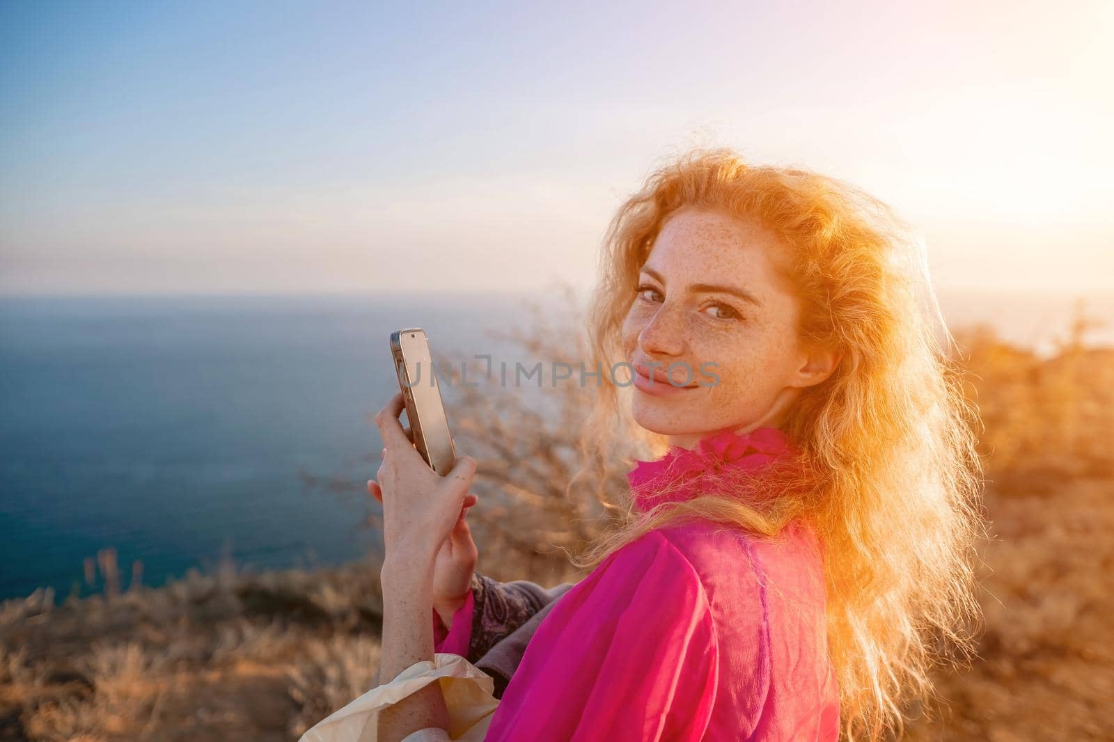 Side view a Young beautiful sensual woman in a red long dress posing on a volcanic rock high above the sea during sunset. Girl on the nature on overcast sky background. Fashion photo