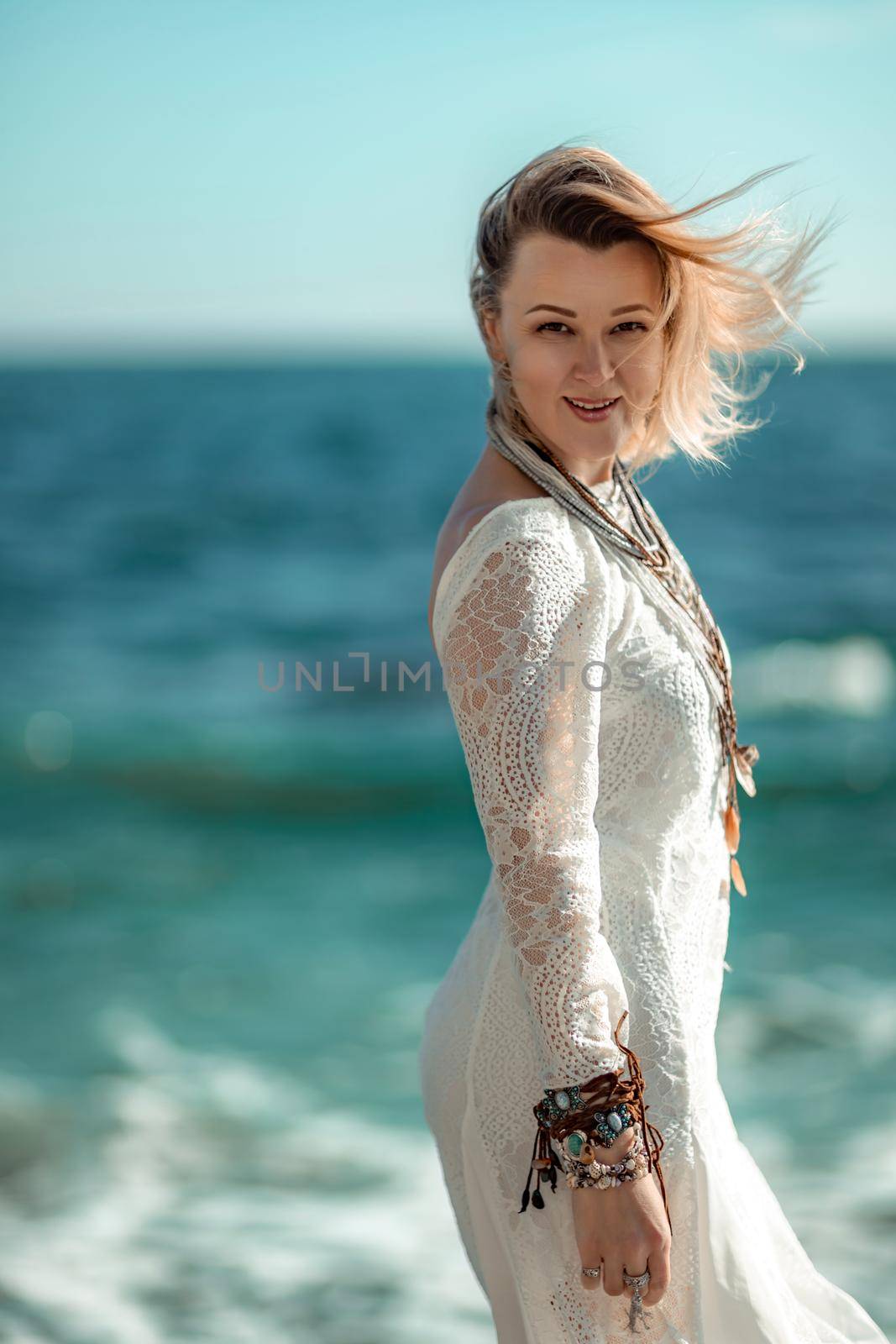 Middle aged woman looks good with blond hair, boho style in white long dress on the beach decorations on her neck and arms. by Matiunina