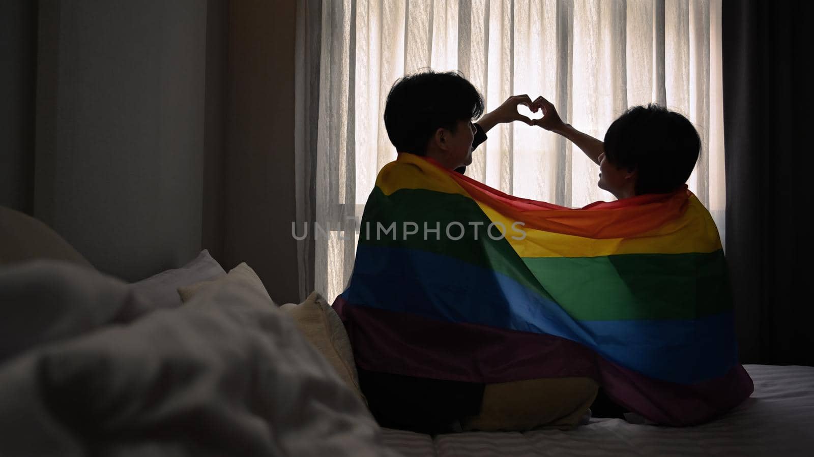 Happy same sex male couple making heart with their hands while sitting under rainbow flag. LGBT, pride, relationships and equality concept.