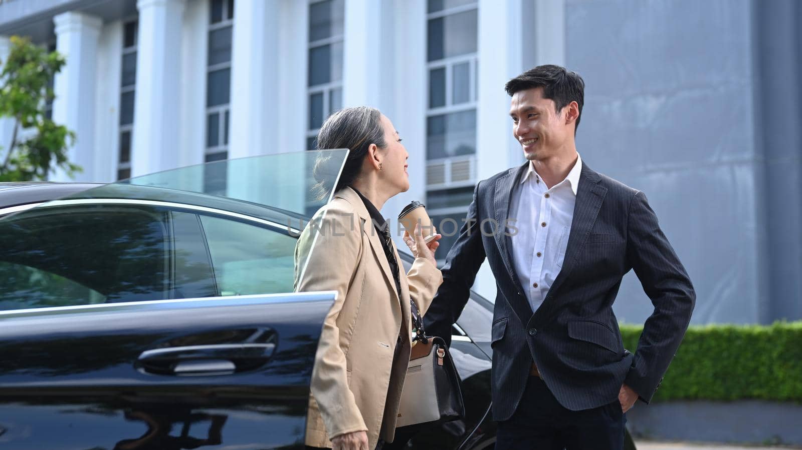 Senior business woman discussing something with young businessman while standing outdoors near a car by prathanchorruangsak