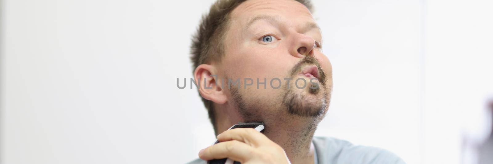 Portrait of middle aged man shaving beard with machine. Electronic device for quick face care, get ready in morning, look in mirror. Good morning, bathroom, beauty treatment concept