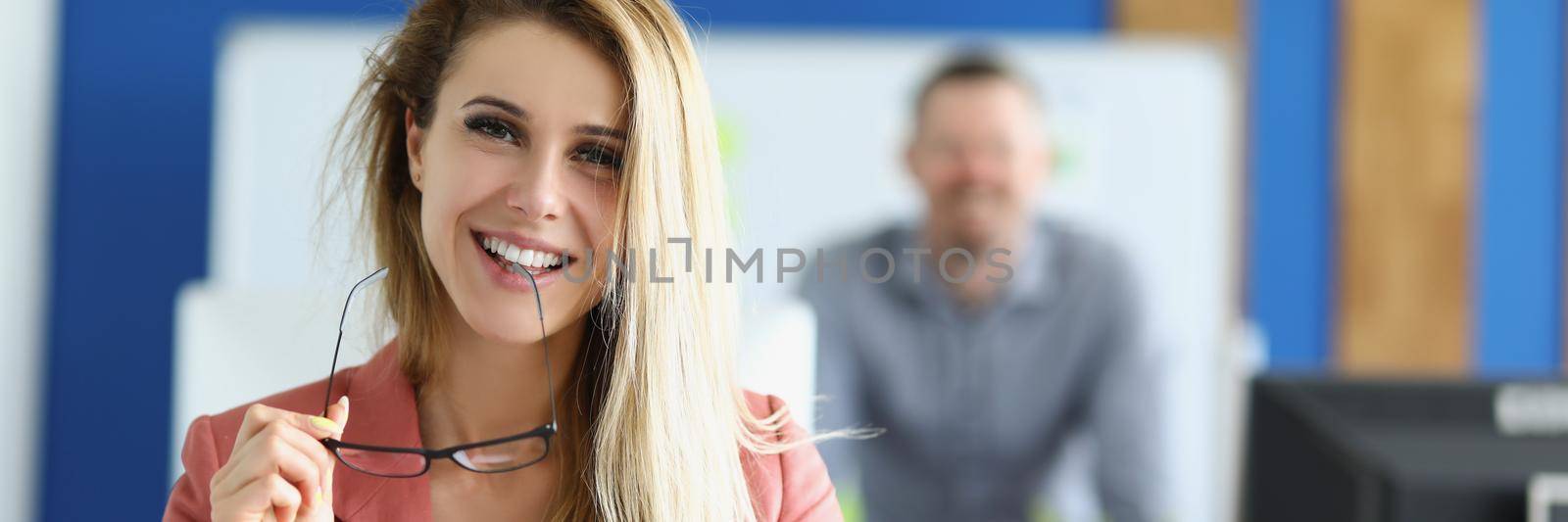 Portrait of cheerful optimistic female with clipboard, company dress code, office worker in suit. Perform daily tasks on workplace, woman employee, colleague on background. Business, career concept