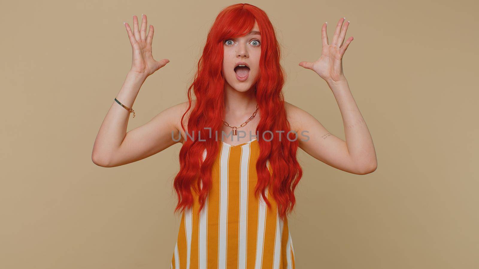 Oh my God, Wow. Excited amazed winner ginger woman raising hands in surprise looking at camera with big eyes, shocked by sudden victory. Sincere young girl isolated alone on beige studio background
